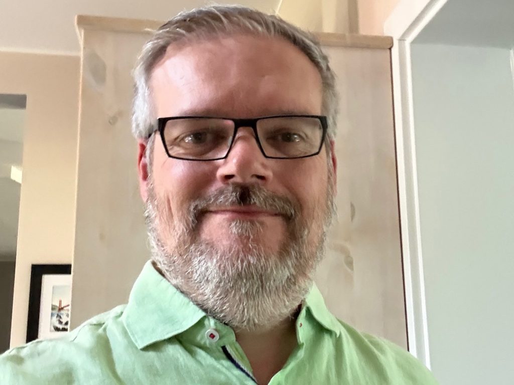Photograph of Marc Zimer; a headshot of a white man with gray hair, a beard and glasses wearing a light green shirt.