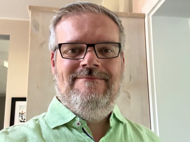 Photograph of Marc Zimer; a headshot of a white man with gray hair, a beard and glasses wearing a light green shirt.